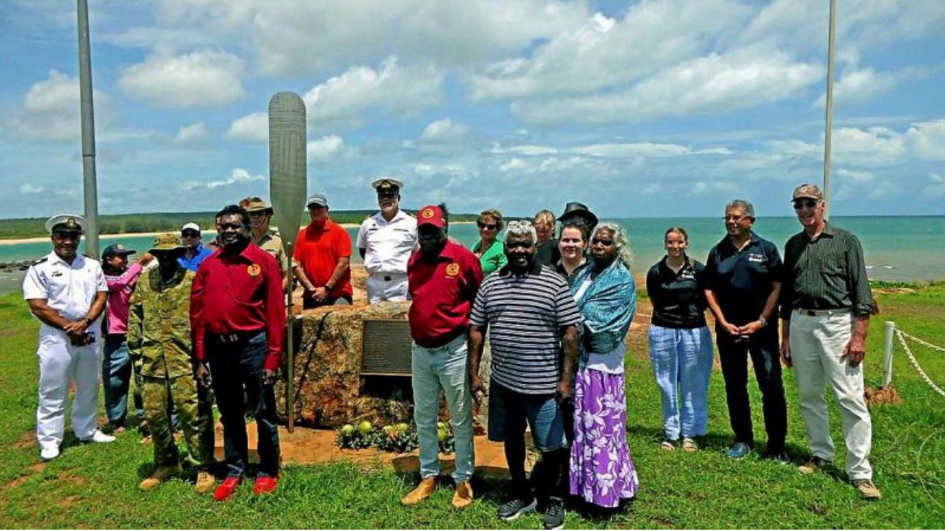 About 20 people stand next to memorial on tropical coastline