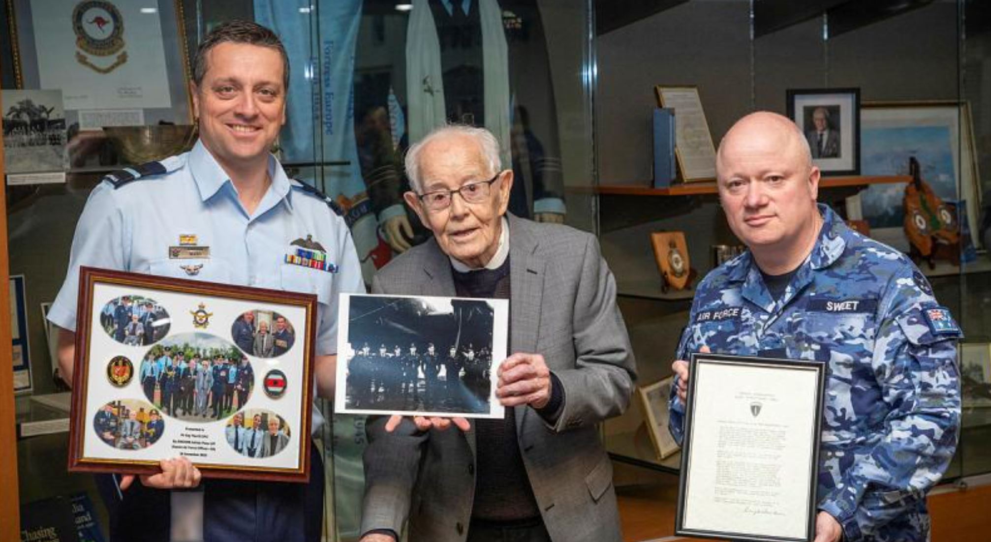 An old man and two younger men in Air Force uniforms hold frames photos and a letter