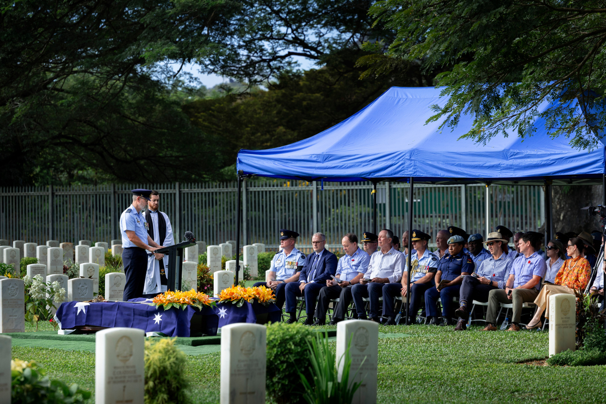 RAAF officer stands at lectern in front of dozens of people under marquee in cemetery.