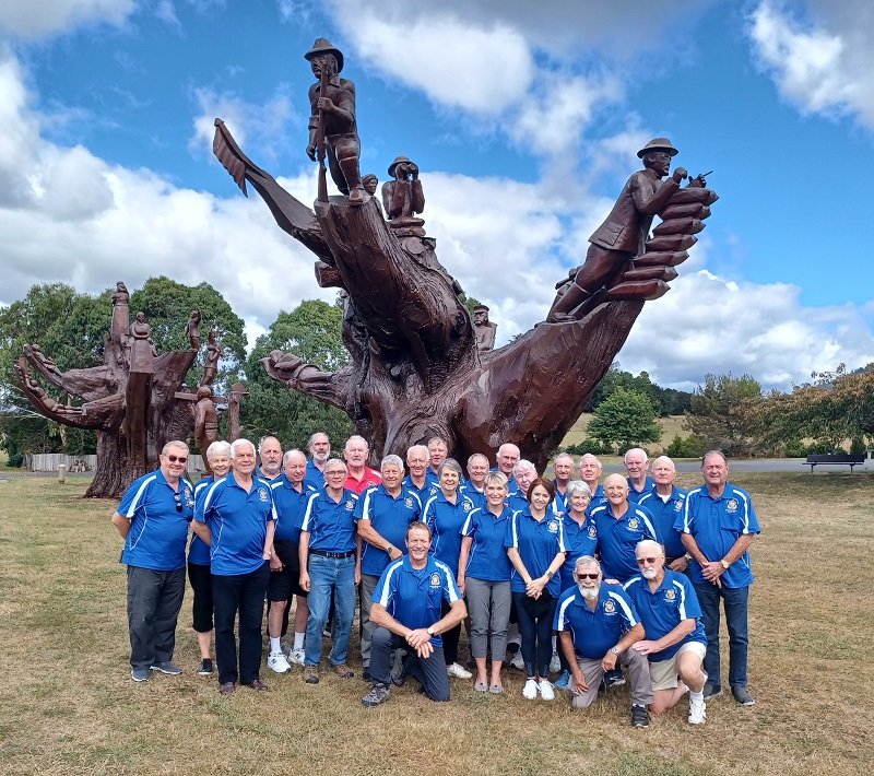 A large group of men and women wearing blue T-shirts pose in front of large sculpted tree stump