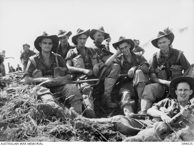 Seven smiling soldiers pose for photo while sitting on rough ground