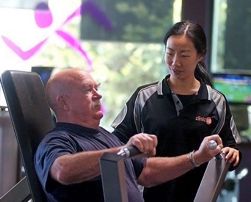 A young female exercise physiologist (EP) or physiotherapist assisting an elderly male patient in a gym.