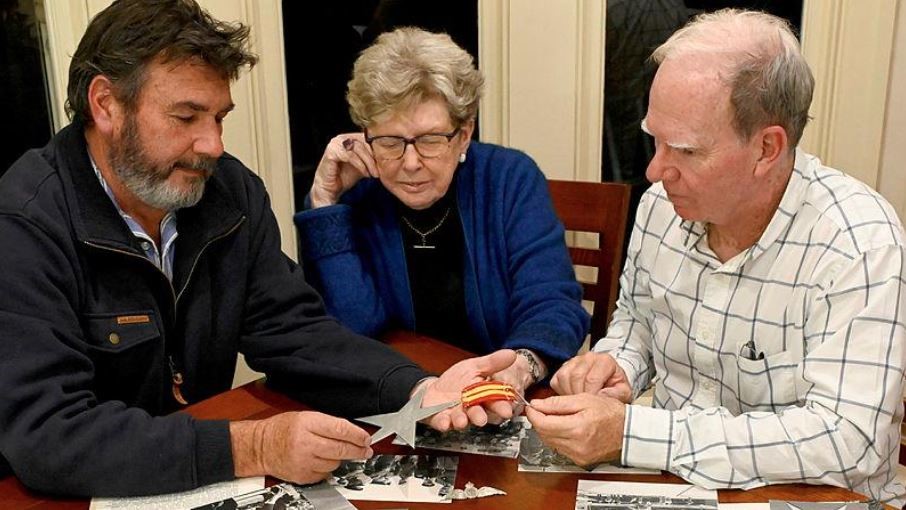 Two middle-aged men and a middle-aged woman look at medals on a table