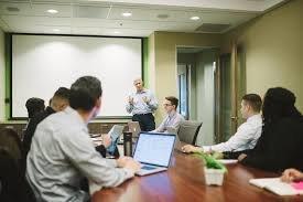 A group of people are sitting around a table. The people have laptops open in front of them. A man is standing at the head of the table, speaking. 