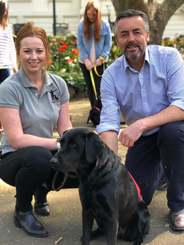 A man and woman crouched behind a black coloured labrador dog