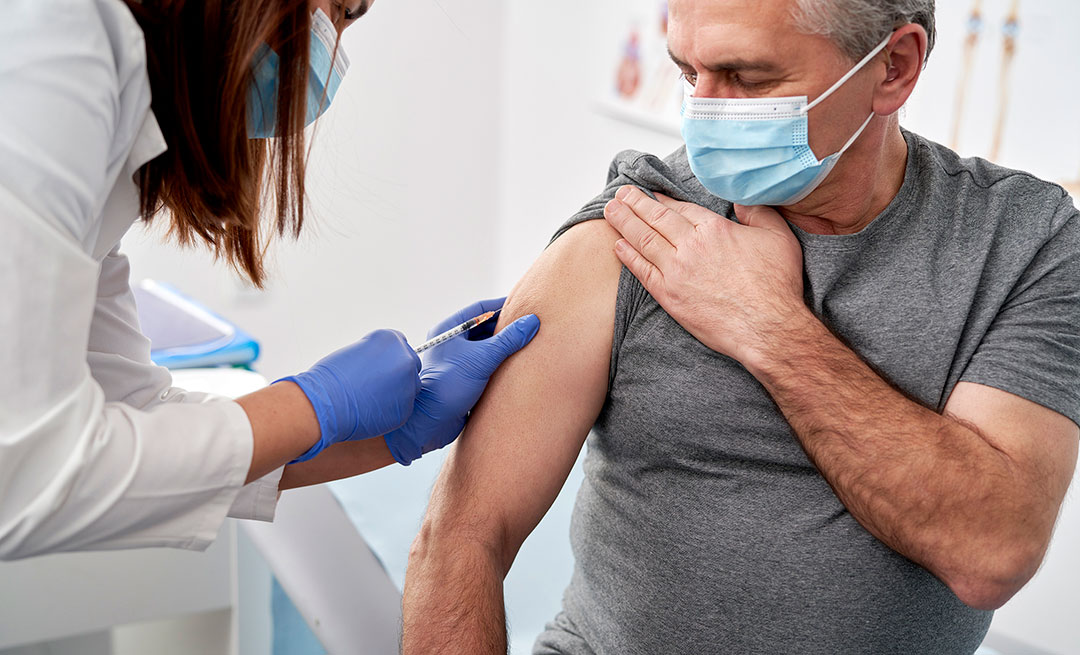 A woman, with long brown hair and wearing a white, medical lab coat, blue gloves and light blue mask. She is injecting a needle into the right arm of an elderly man. The man is sitting down and wearing a grey t-shirt and a light blue mask