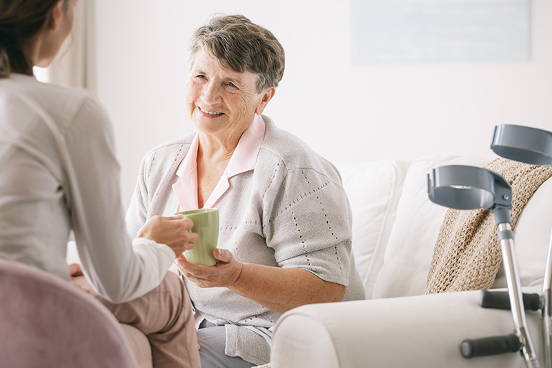 An older lady with her happy smile is having a cup of drink with another younger lady. There are a pair of walking crutches next to them. For  thickened fluid image