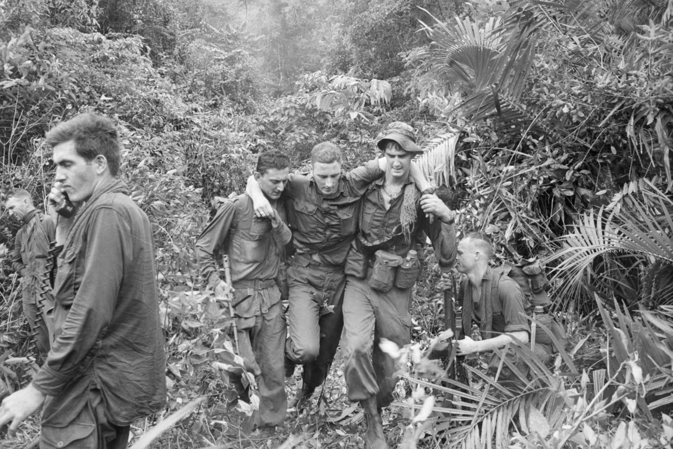 A wounded National Serviceman from 5RAR being helped to a helicopter evacuation site in Phuoc Tuy Province, Vietnam (AWM COA/66/0877/N).