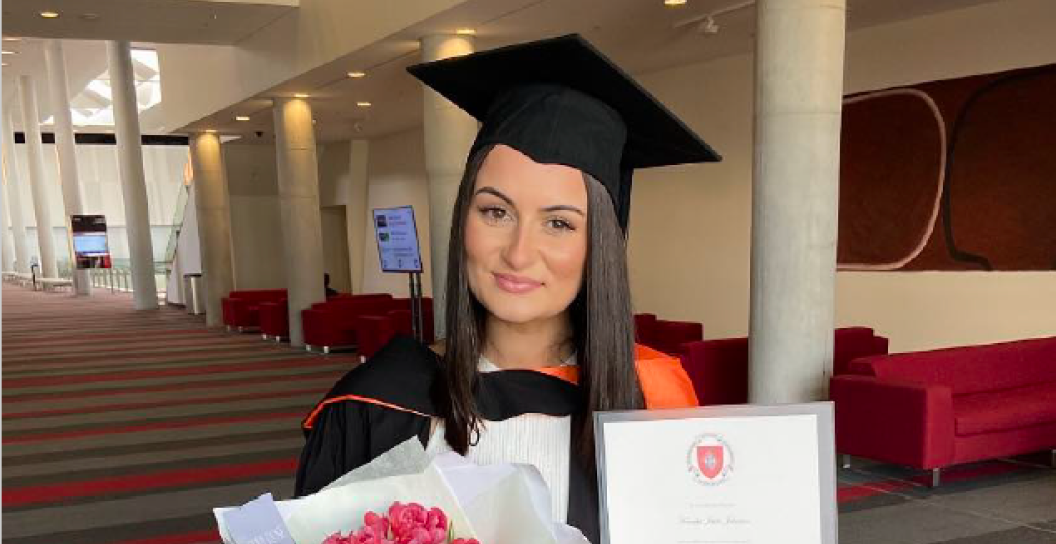 An image of a young woman on university graduation day. She is smiling while holding red flowers and her degree.