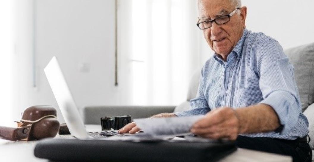 Elderly gentleman wearing glasses, reading piece of paper while sitting in front of a laptop