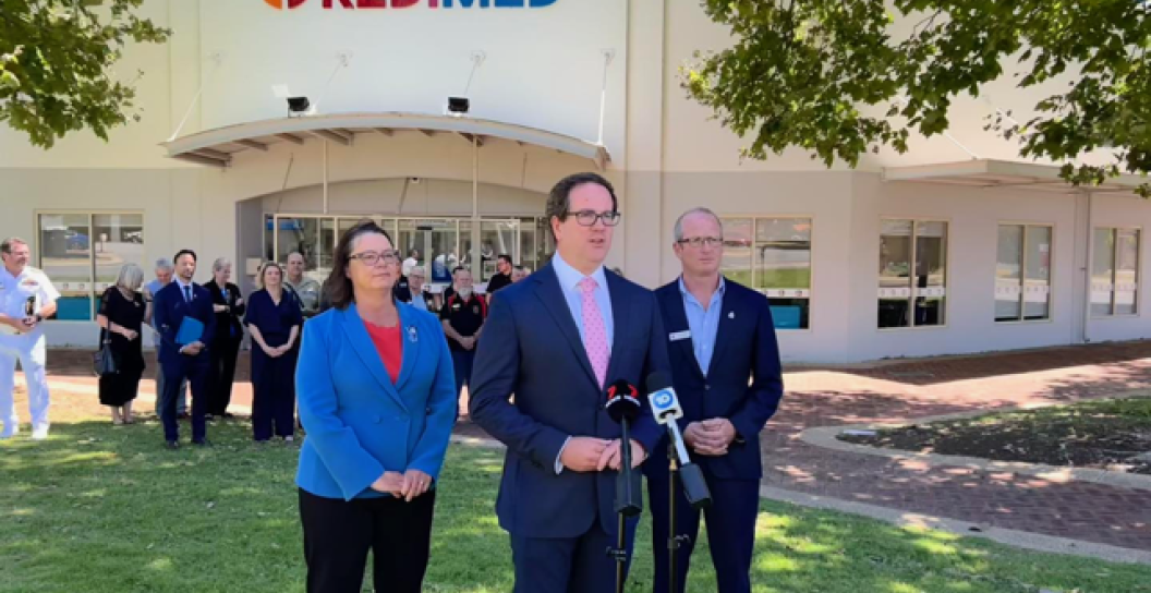 Three people standing in front of a microphone in front of a white building; a small crowd stand behind them.