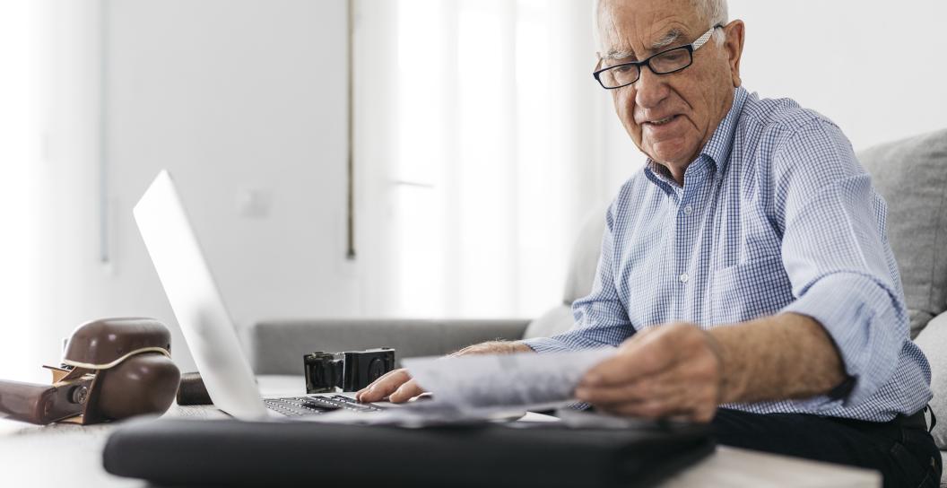Elderly gentleman wearing glasses, reading piece of paper while sitting in front of a laptop