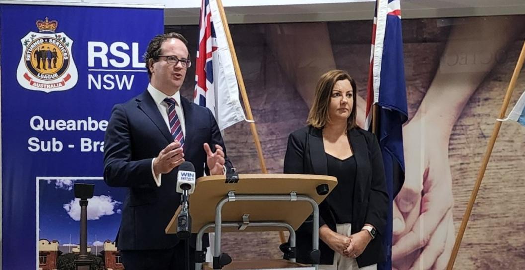 Man and a woman behind lectern, with RSL sign and two flags behind them.