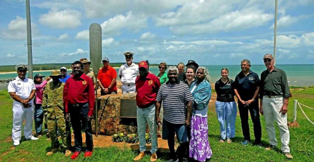 About 20 people stand next to memorial on tropical coastline