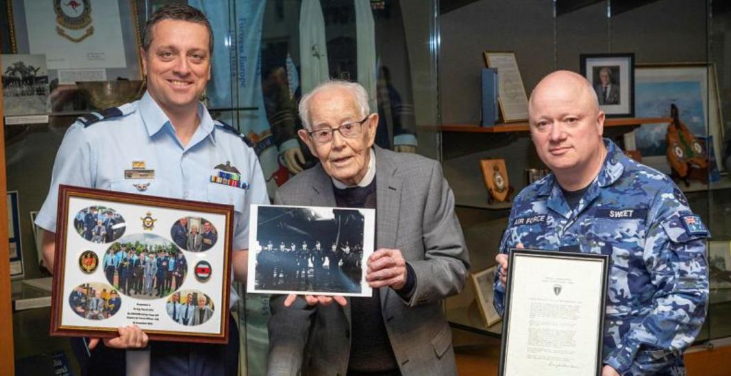 An old man and two younger men in Air Force uniforms hold frames photos and a letter