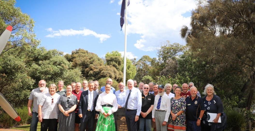Group of some 20 people standing in front of a flag pole with trees in the background