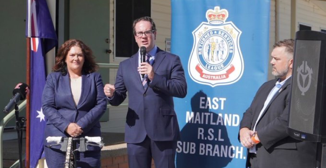 Man speaking into microphone, with a person on either side, in front of RSL banner