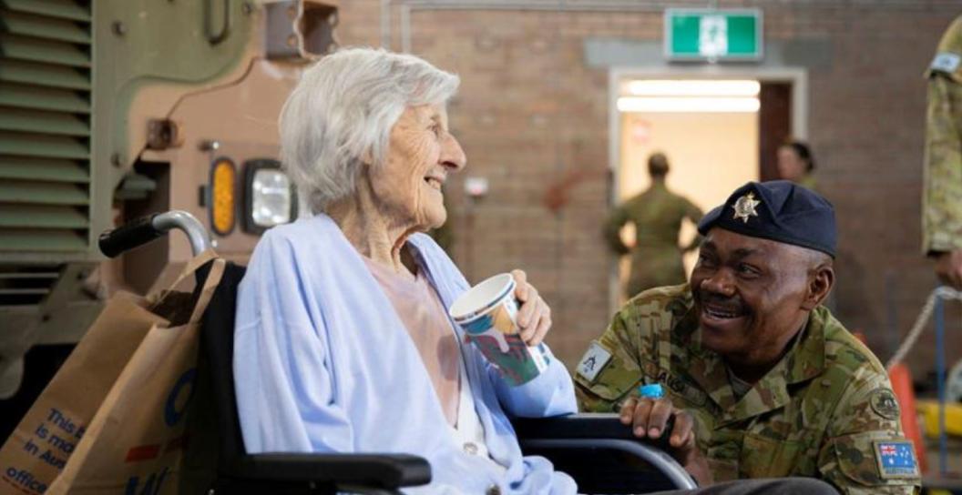 An elderly lady in wheelchair next to Army truck chats with soldier