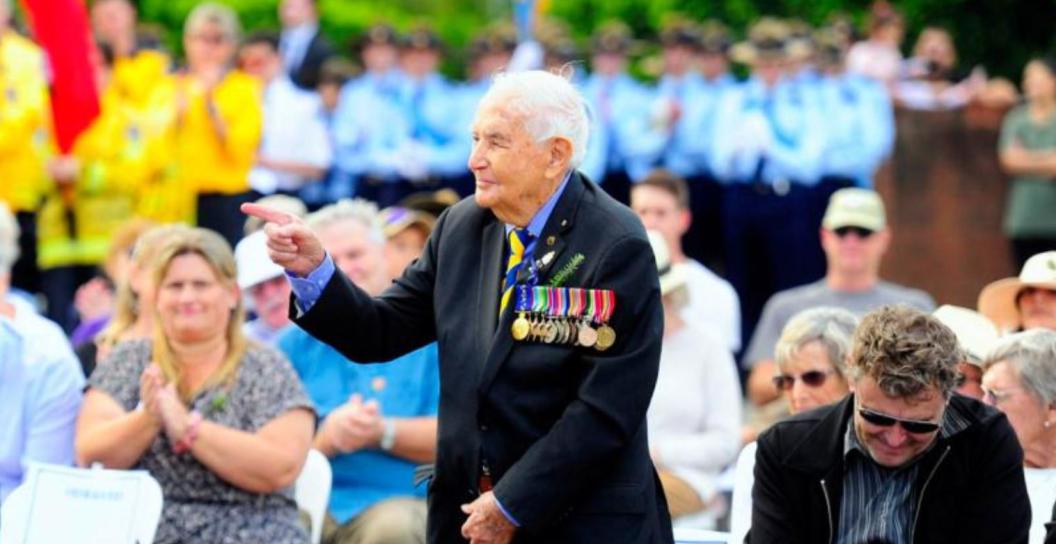 Older man wearing medals standing up in front of applauding crowd