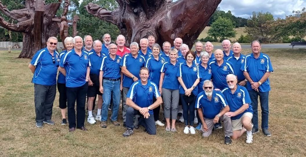 Twenty or so older men and one woman in blue T-shirts pose in front of large sculpted tree stump
