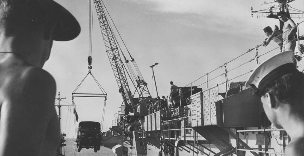 A soldier and a sailor watch a jeep being loaded or unloaded onto an aircraft carrier