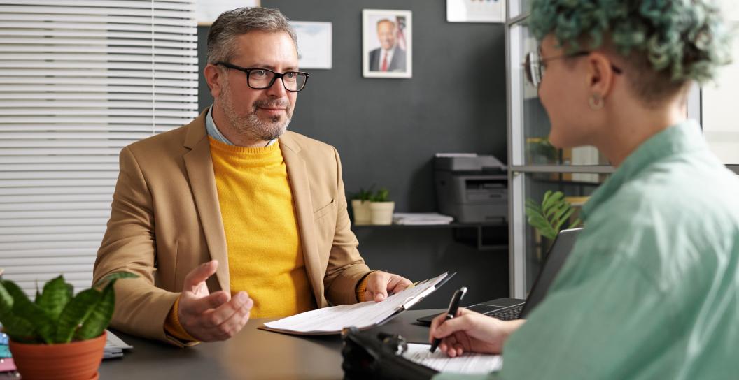 woman with black prosthetic arm signing documents at desk across from man in yellow shirt
