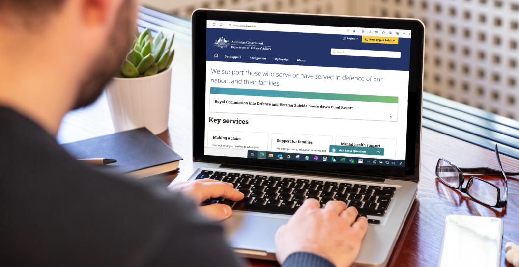 Man using a laptop at a desk. The laptop screen shows the DVA website.