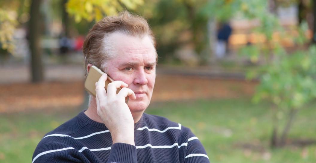 A man in his 40s or 50s is sitting on a park bench on a mobile phone call. His brow is furrowed, and he seems suspicious of the person he is talking with.