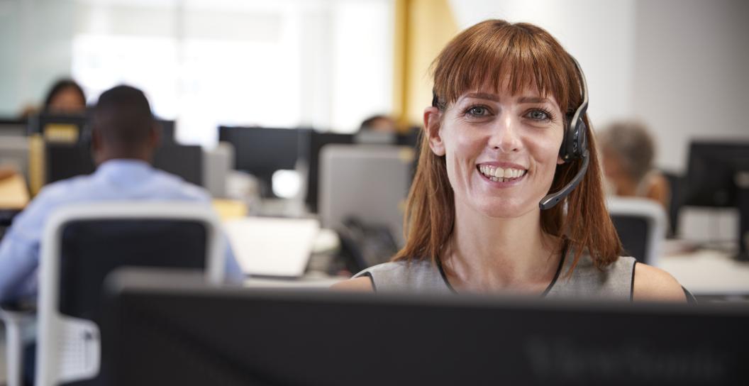 Woman with red hair shoulder length with fringe sits at a computer desk in an office, wearing a headset talking to someone on the phone