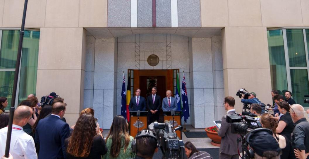 Three men in suits before gaggle of journalists
