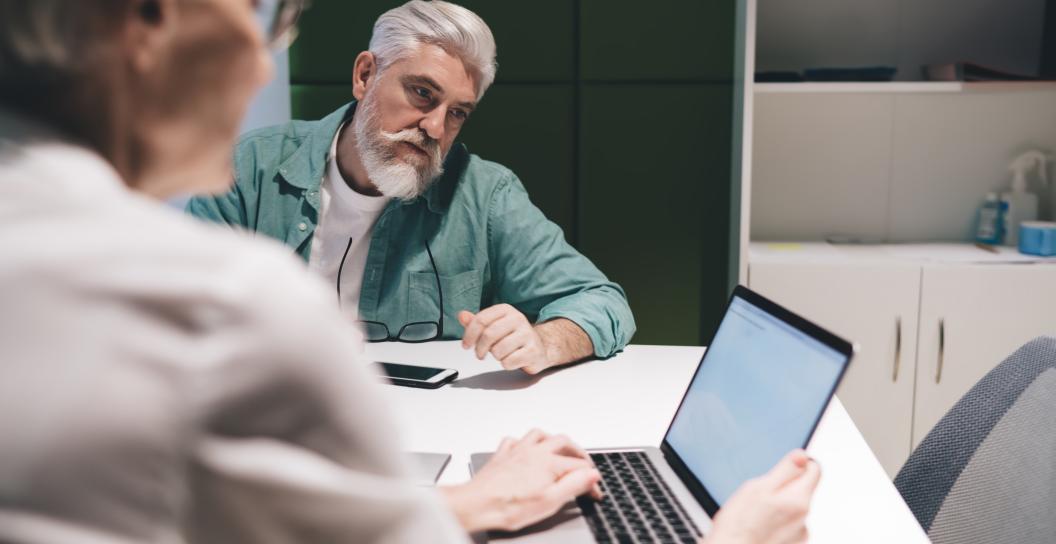 Man talking to woman at desk, with lap top in front of her.