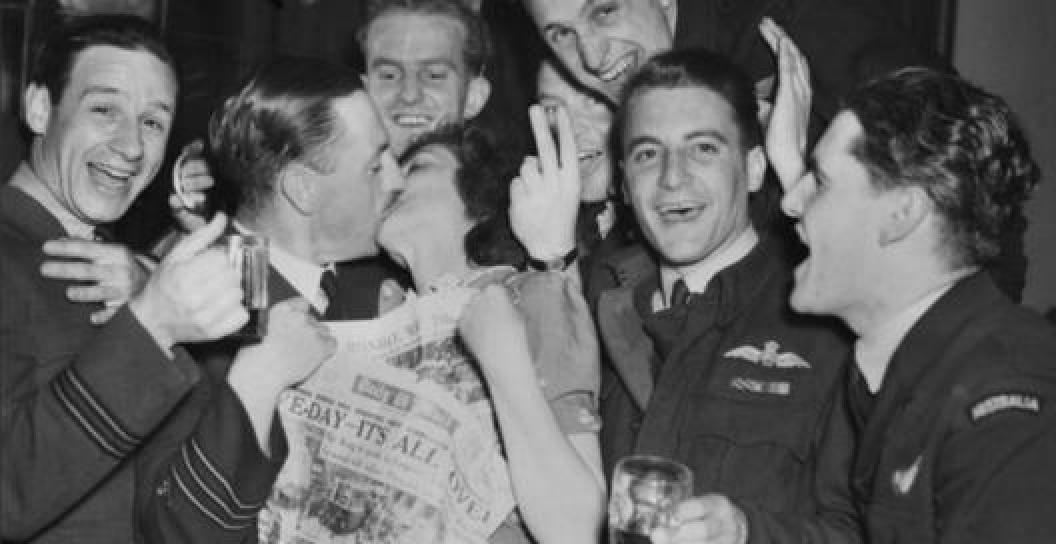 Black and white photo of six happy men in Air Force uniform. One is kissing a woman while holding a newspaper declaring the war is over.