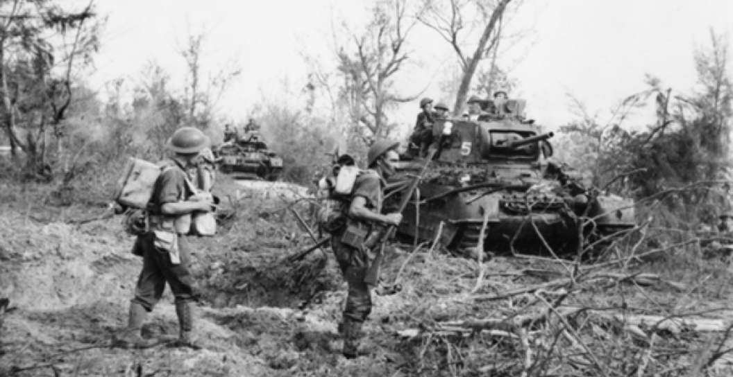 Two soldiers walking in light scrub with a tank in the background