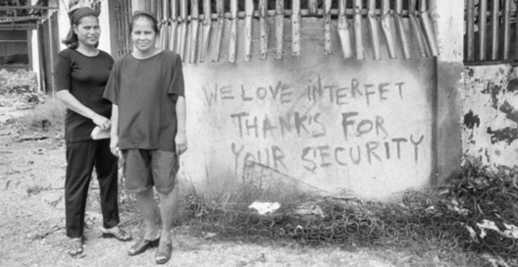 Two East Timorese women stand beside a hand painted slogan on the wall of a derelict building. The sign reads 'We love INTERFET. Thanks for your security'.