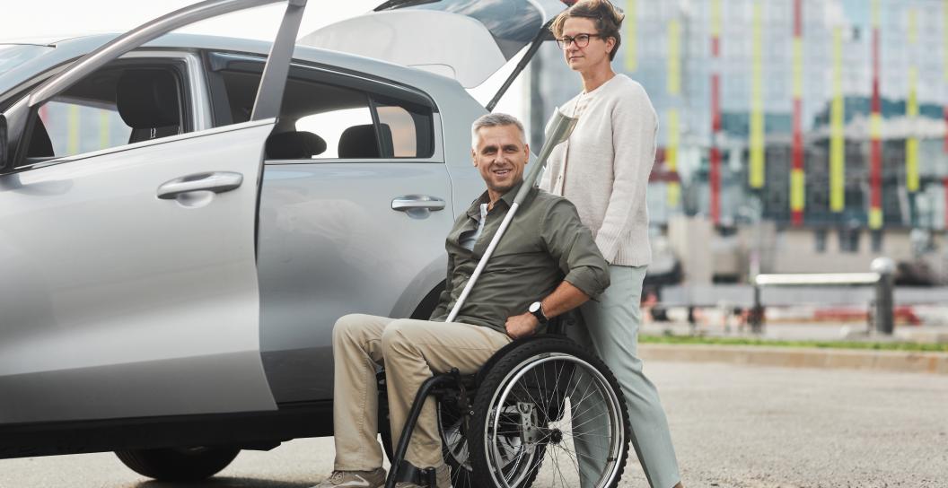 Man in wheelchair is being put into a car by his female carer outside a hospital