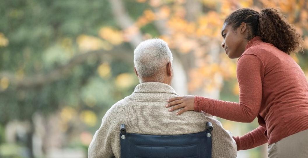 Man in a wheelchair in a park. Woman has her hand on his back. 