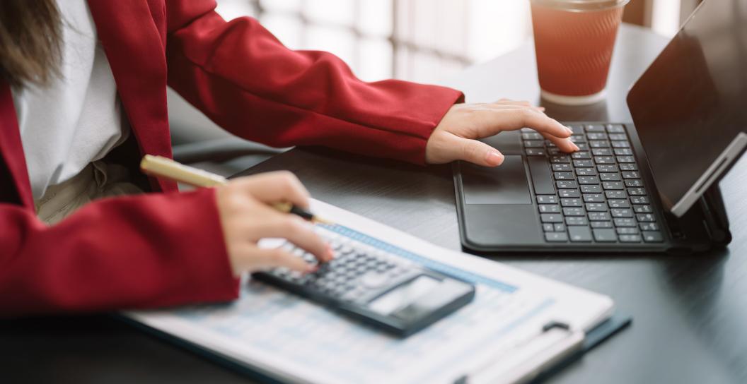A person, whose gender is unknown, working at a desk with a laptop, calculator, pen, and a drink. The person is wearing a red blazer.