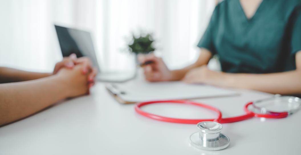 A health professional, wearing a dark green top, sits at a white desk with a patient. A red stethoscope lies on the desk in the foreground, along with a clipboard, pen, laptop, and decorative plant. 